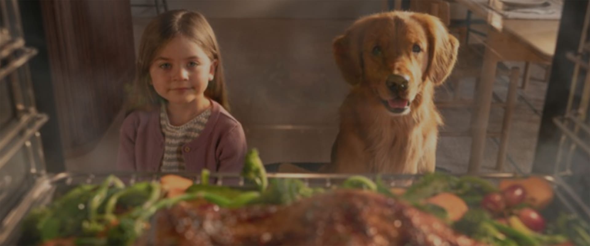 A girl and a dog are looking at the food being cooked in an oven.